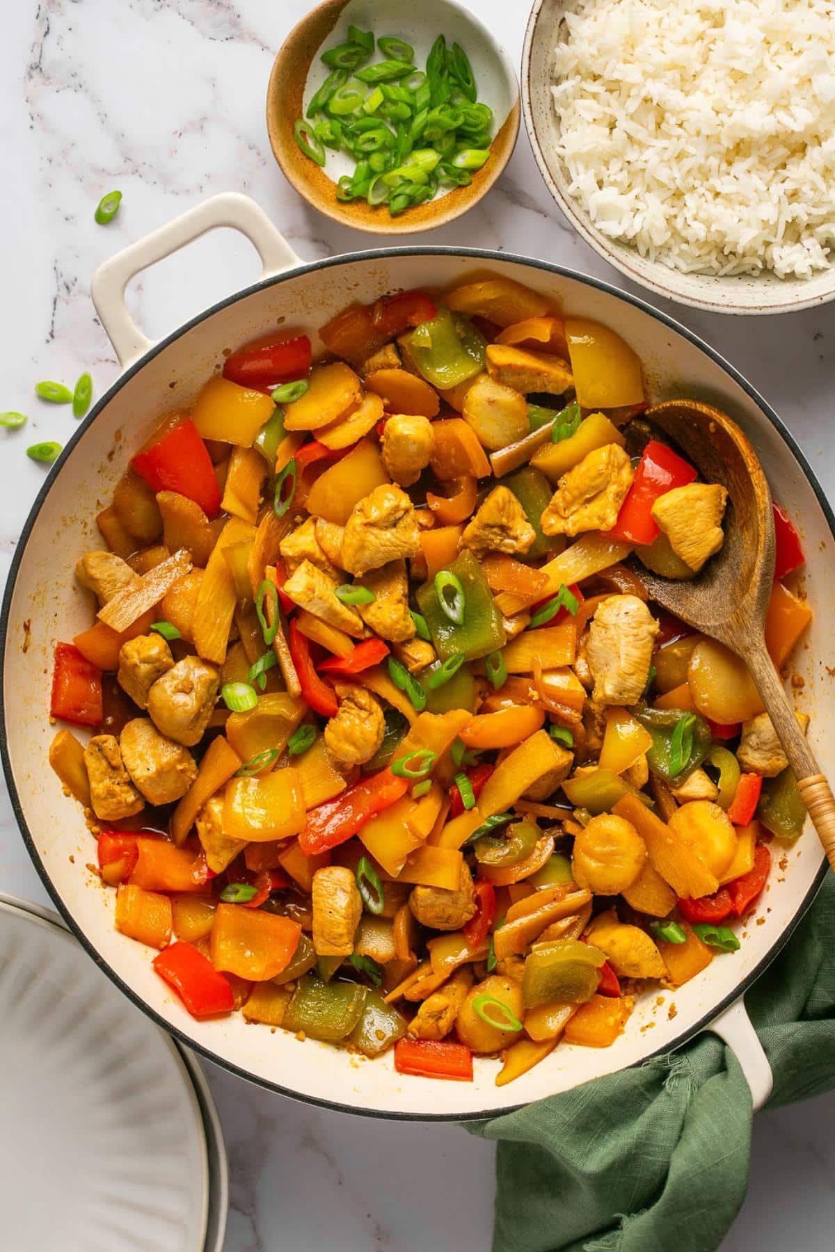 A healthy chicken stir fry in a skillet with a wooden spoon and a dish of green onions and rice to the side.