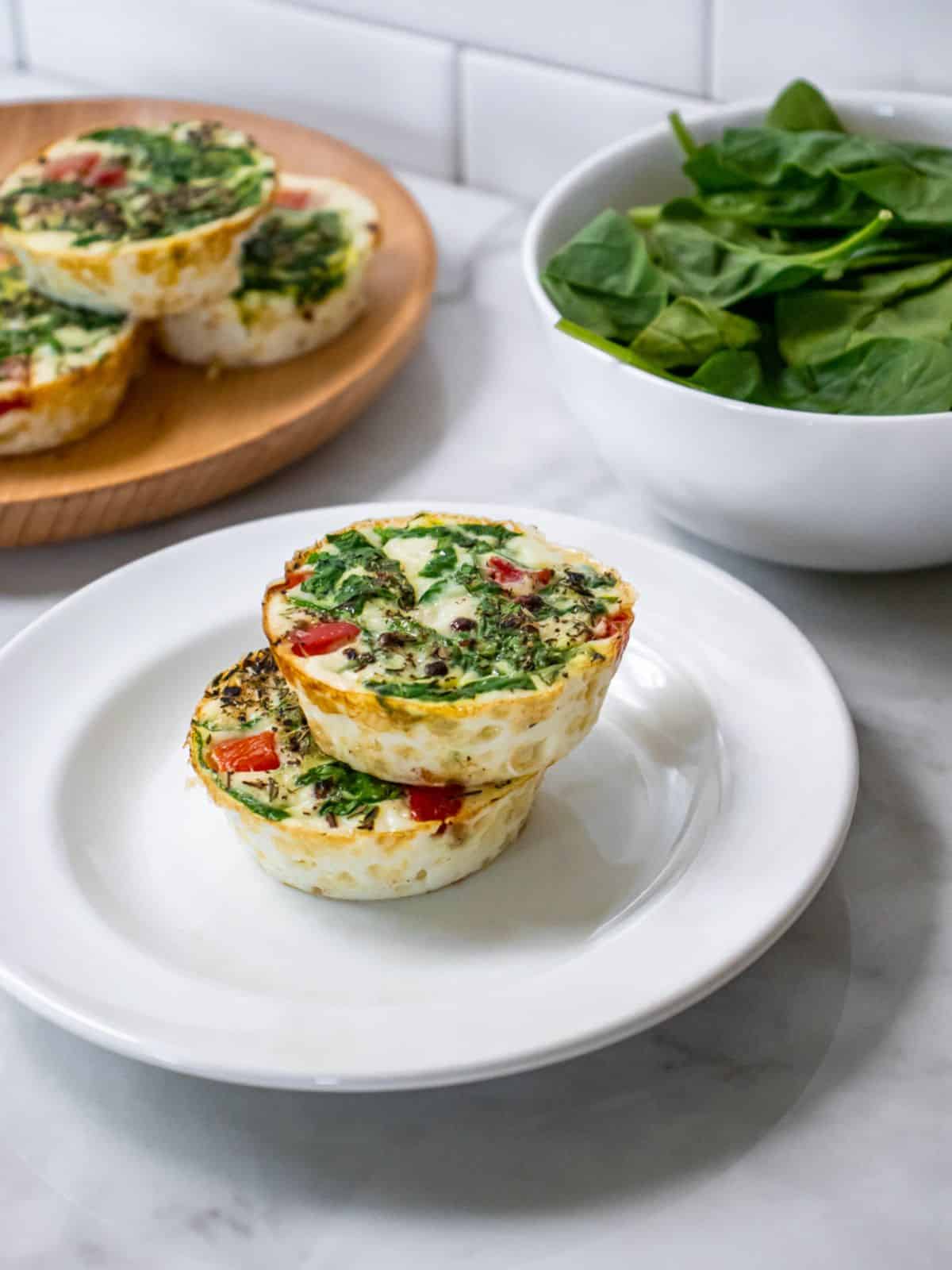 Two egg white egg bites on a white plate with a platter of more in the background.