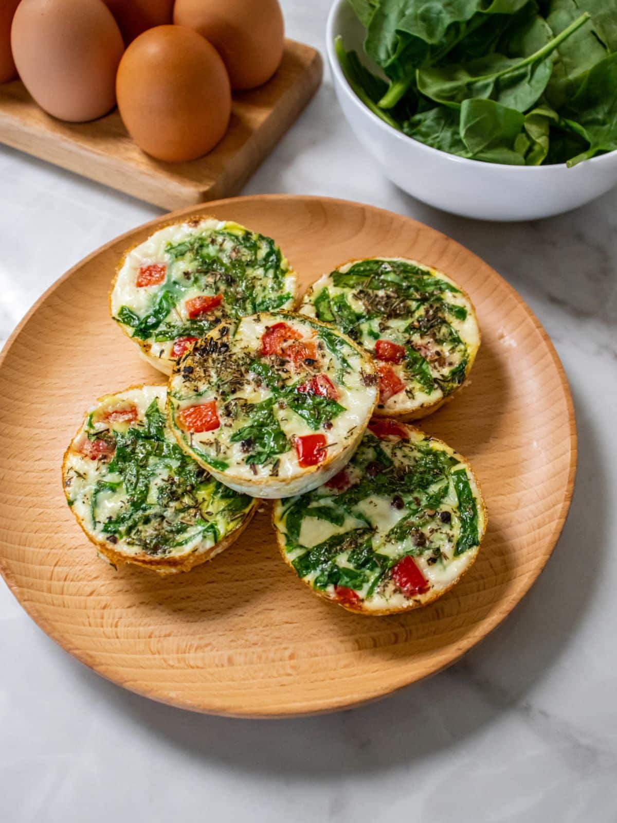 Egg white bites with spinach and red pepper on a round wooden plate with eggs and fresh spinach in the background.
