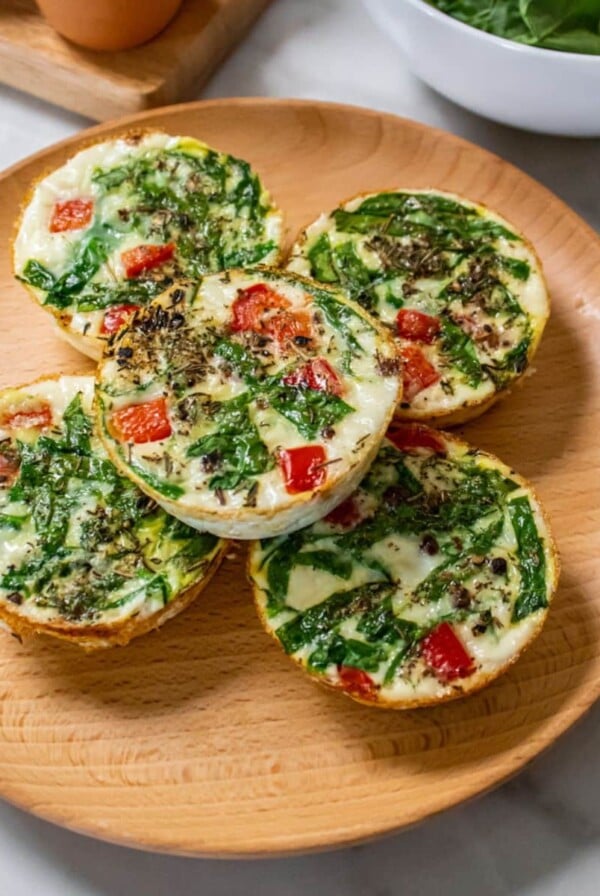 Egg white bites on a wooden plate with a bowl of spinach and whole eggs in the background.