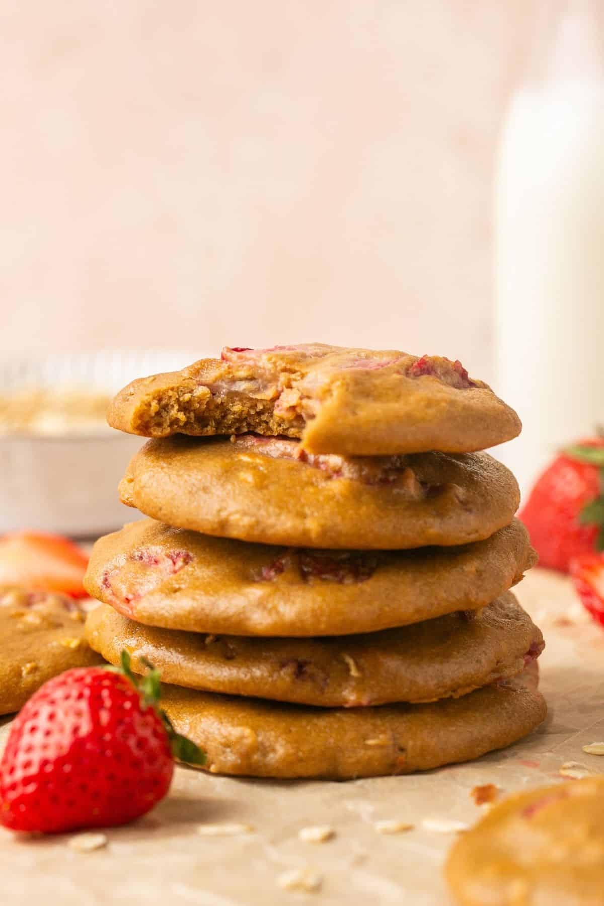 A stack of strawberry oatmeal cookies with a bottle of milk in the background and fresh berries scattered around.