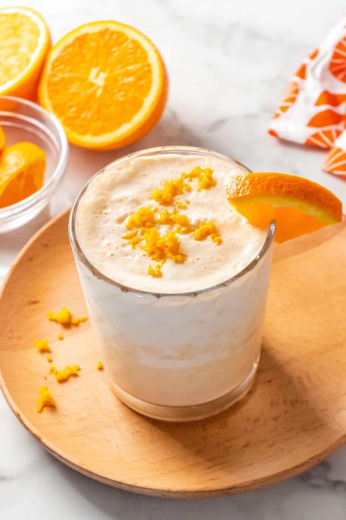 A healthy orange julius in a glass topped with some orange zest and an orange piece on the glass with more oranges in the background.