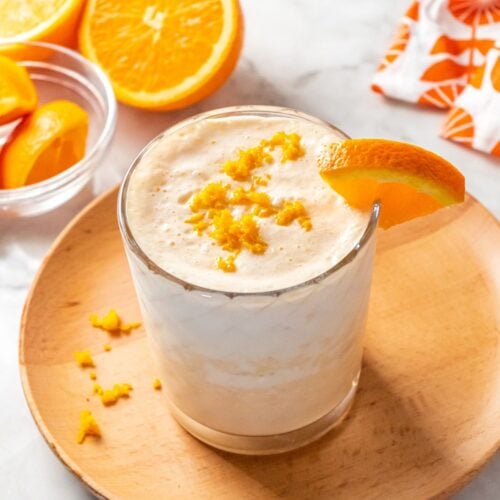 A healthy orange smoothie on a small round wooden plate garnished with an orange on the glass and a sprinkle of orange zest.