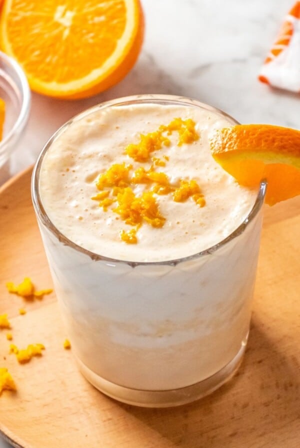 A healthy orange smoothie on a small round wooden plate garnished with an orange on the glass and a sprinkle of orange zest.