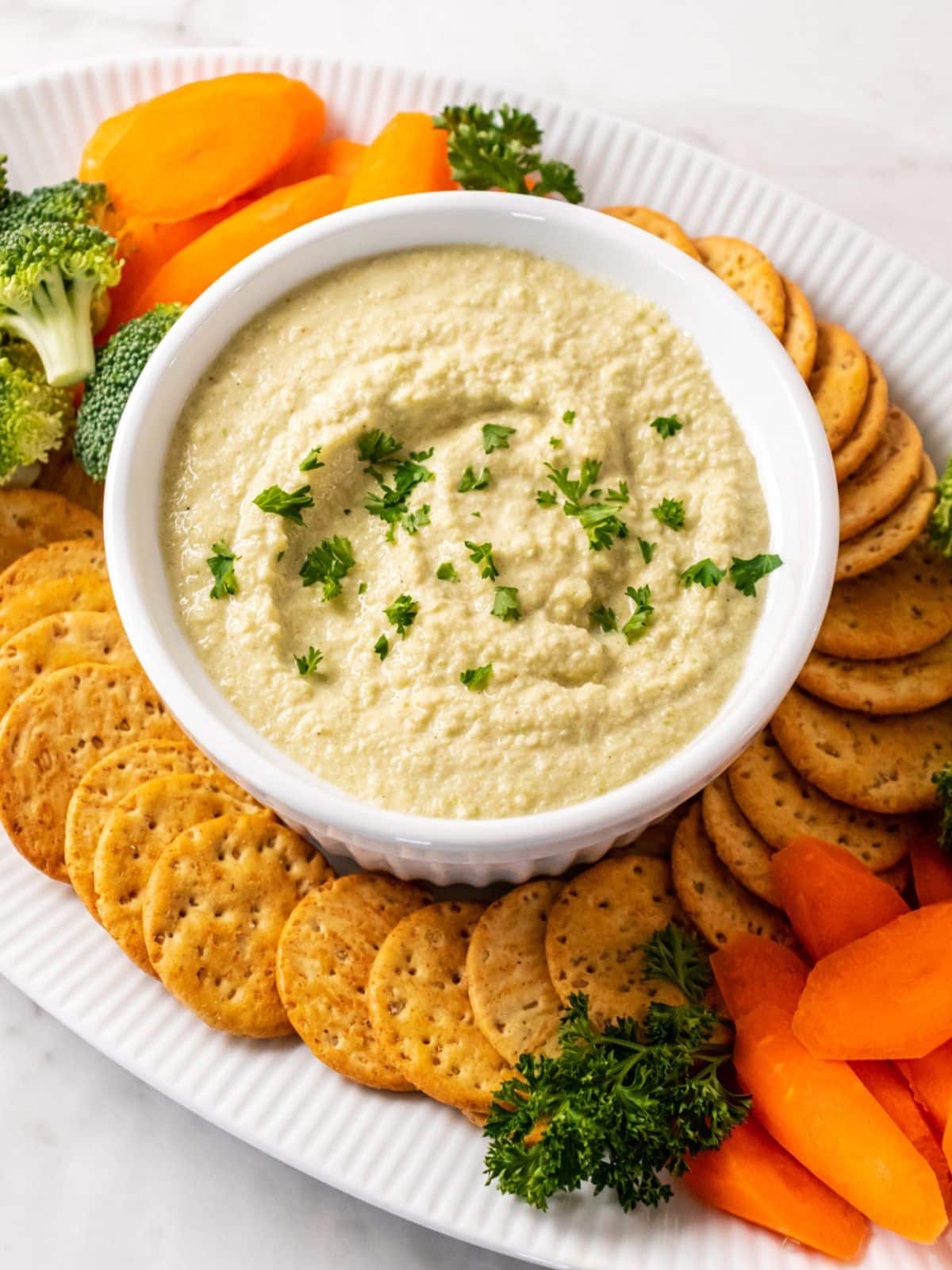 A bowl of zucchini hummus with veggies and crackers on the side.