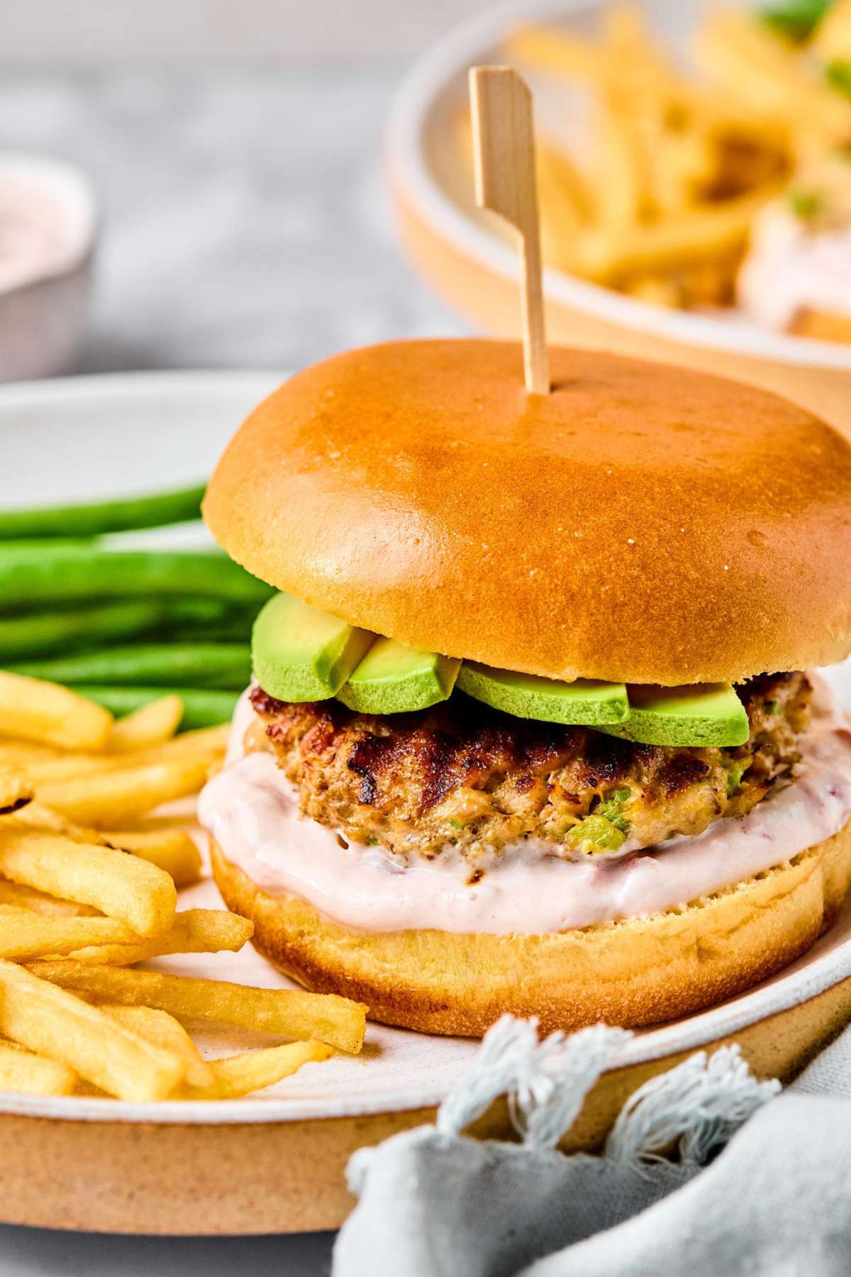 A chicken burger topped with avocado on a plate with fries and green beans.