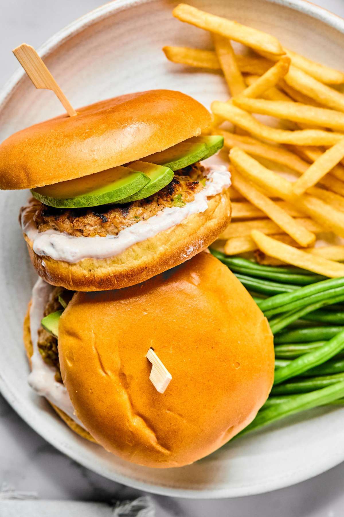Ground chicken burgers on buns with a white sauce and sliced avocado on a plate.