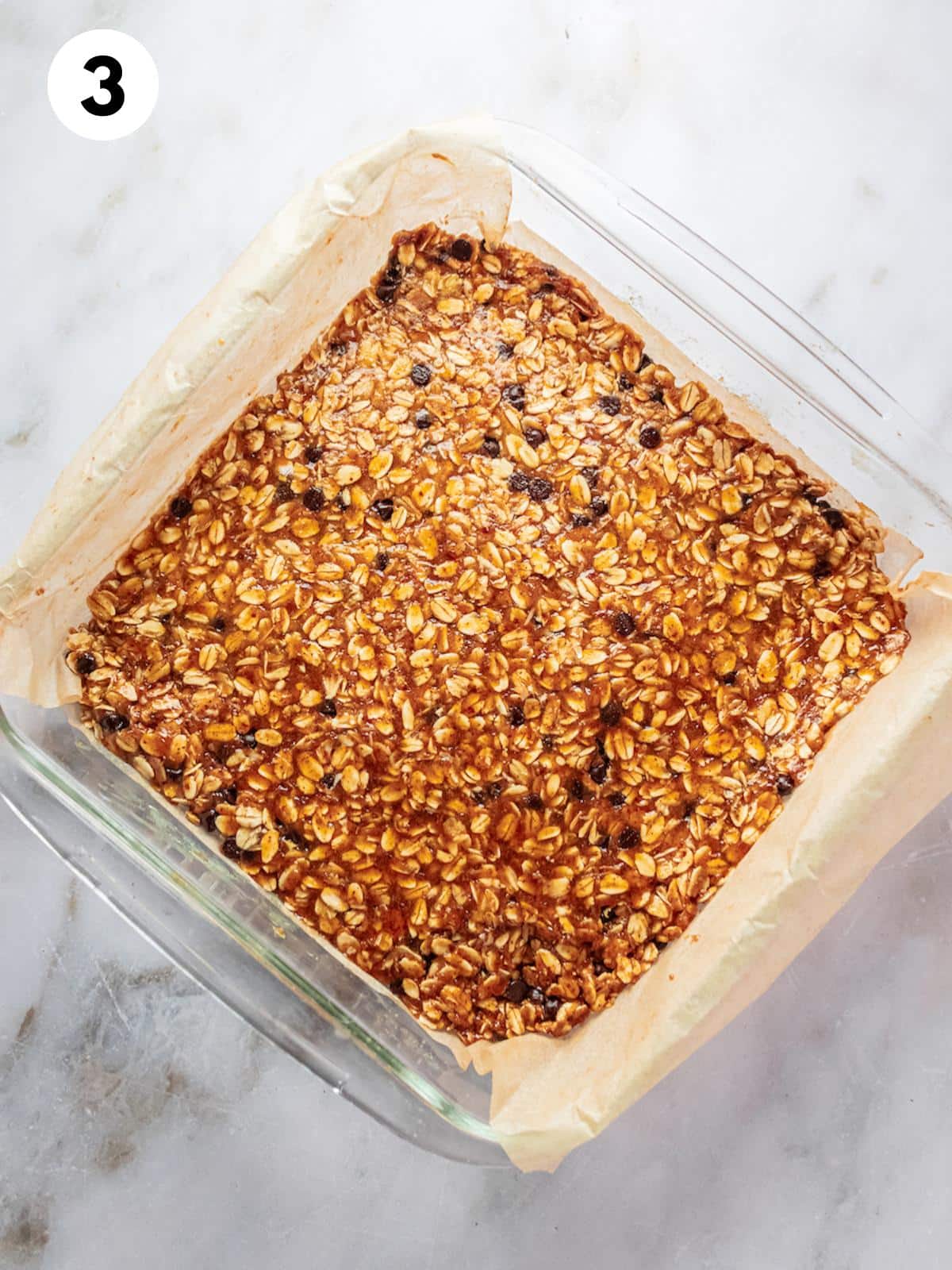 Granola bar mixture pressed into a glass dish lined with parchment paper.