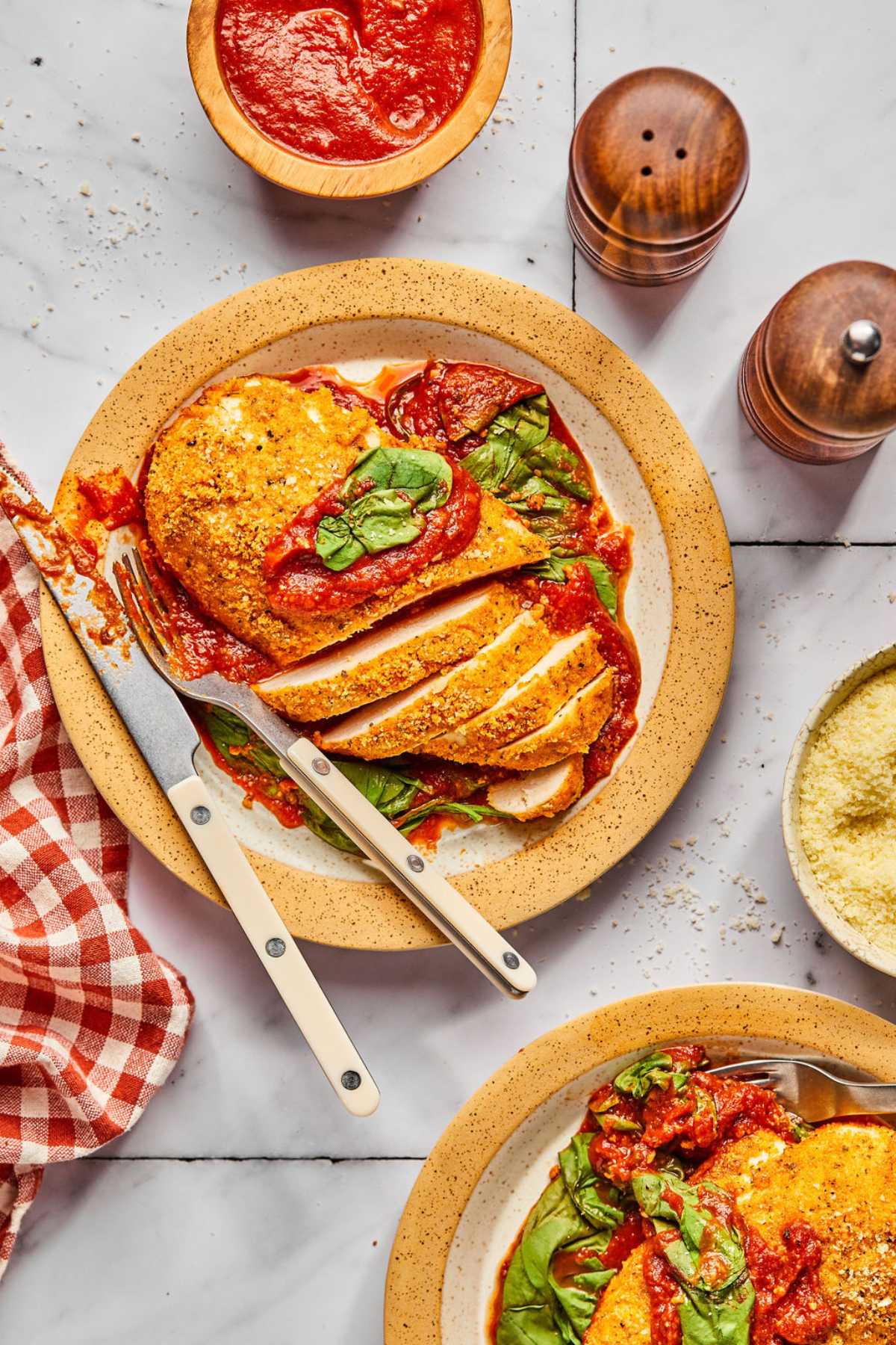 A plate of sheet pan chicken parmesan on the table with half sliced and the fork and knife to the side.