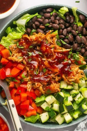 A BBQ chicken bowl on the table with a fork in the bowl and it's topped with chicken, black beans, bell pepper, and zucchini.