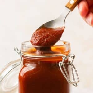 A jar of Low Sugar BBQ Sauce with a hand holding a spoonful over the jar to lift some out.