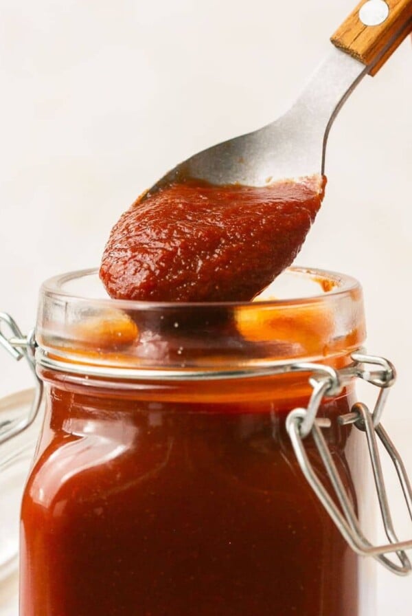 A jar of Low Sugar BBQ Sauce with a hand holding a spoonful over the jar to lift some out.