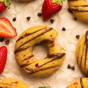 A baked strawberry donut on a piece of parchment with a bite missing and more donuts and berries around it on the paper.