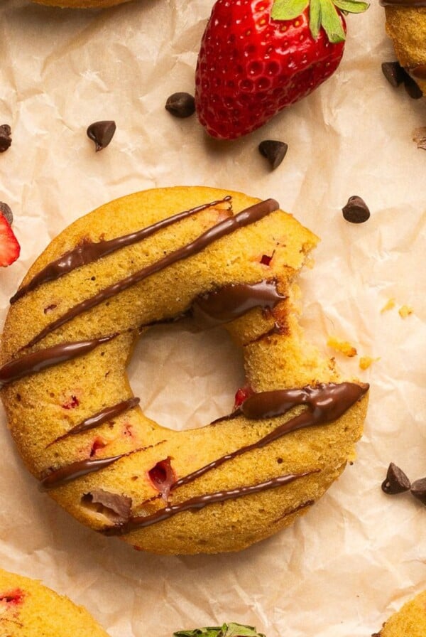 A baked strawberry donut on a piece of parchment with a bite missing and more donuts and berries around it on the paper.