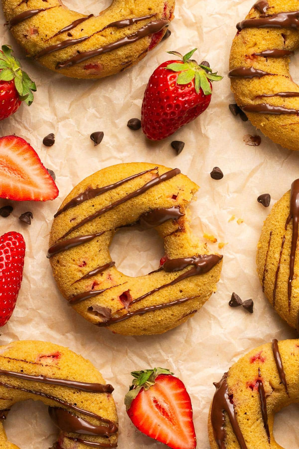 A baked strawberry donut on a piece of parchement with a bite missing and more donuts, berries, and chocolate chips scattered around.