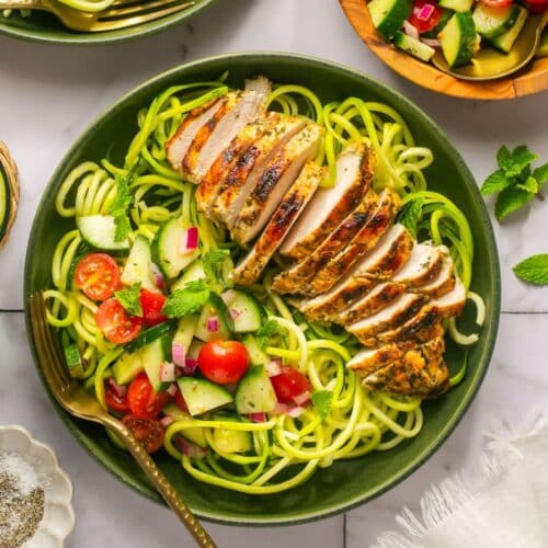 Greek chicken bowls on the table with a a wooden bowl of more salad to the side.