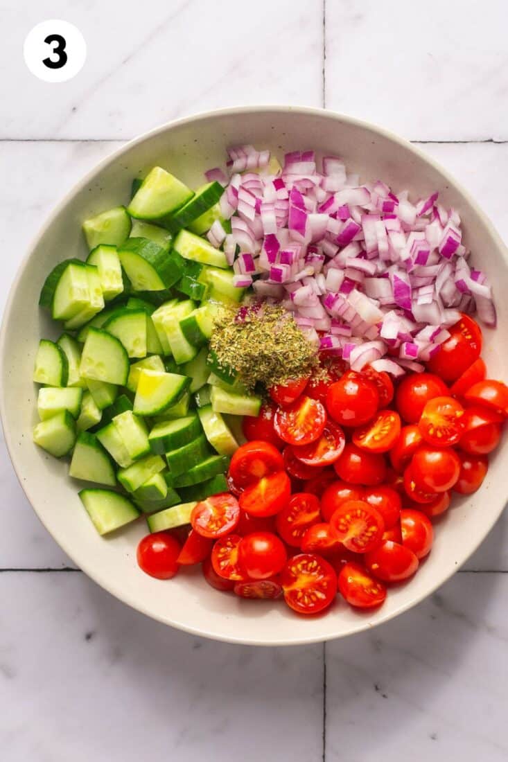 Tomatoes, cucumbers, and red onions in a bowl with seasonings added on top.