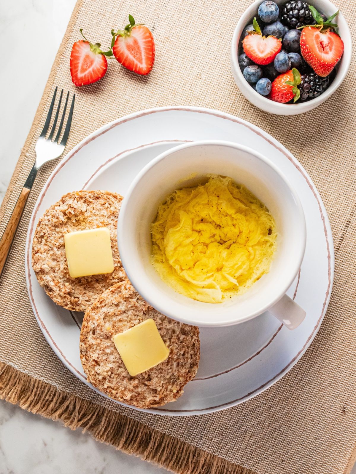 A mug of microwave scrambled eggs on a plate with a bowl of fruit and english muffin.