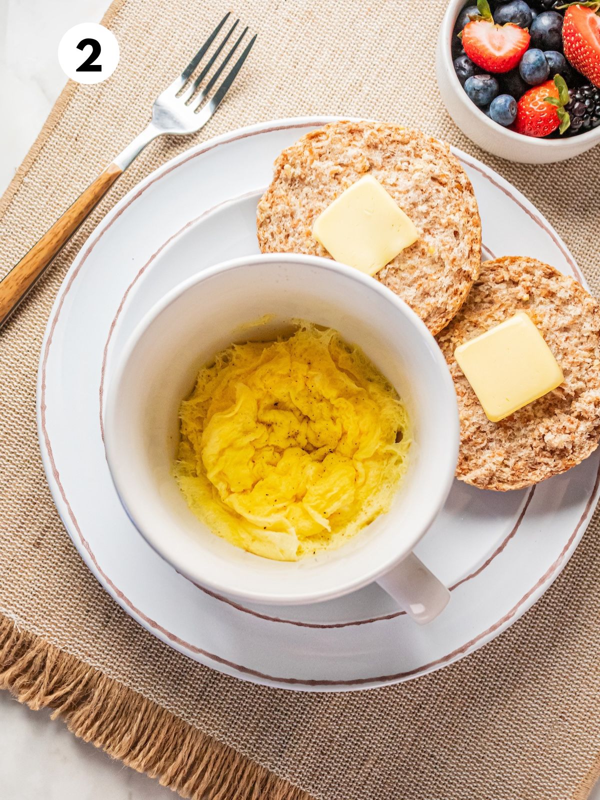 Cooked scrambled eggs in a mug on a plate with an English muffin.
