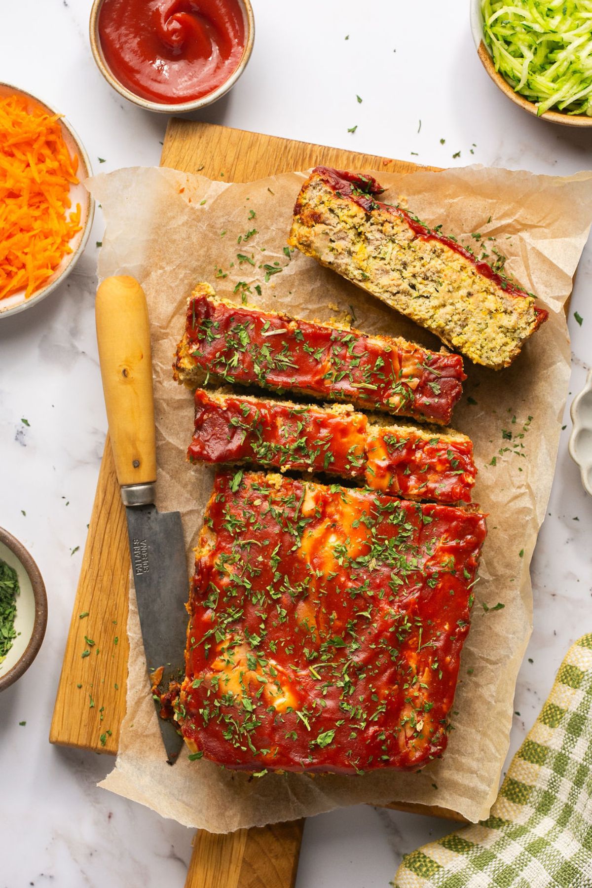 Zucchini meatloaf on the table cut into slices and surrounded by bowls of ingredients on the table. 