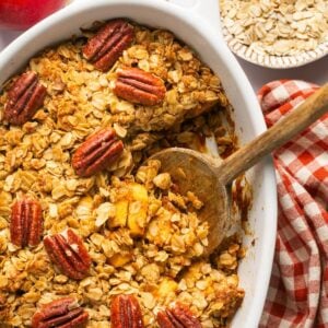 Pumpkin apple crisp topped with pecans in a baking dish with a wooden spoon lifting a serving out.