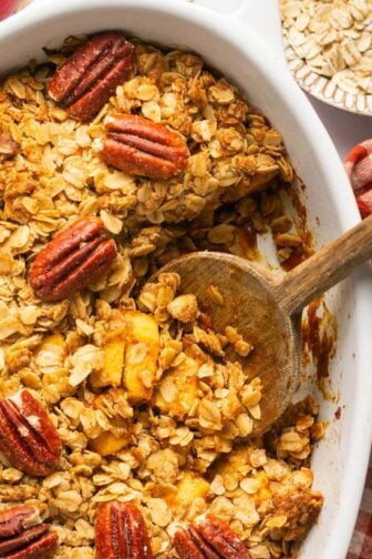 Pumpkin apple crisp topped with pecans in a baking dish with a wooden spoon lifting a serving out.