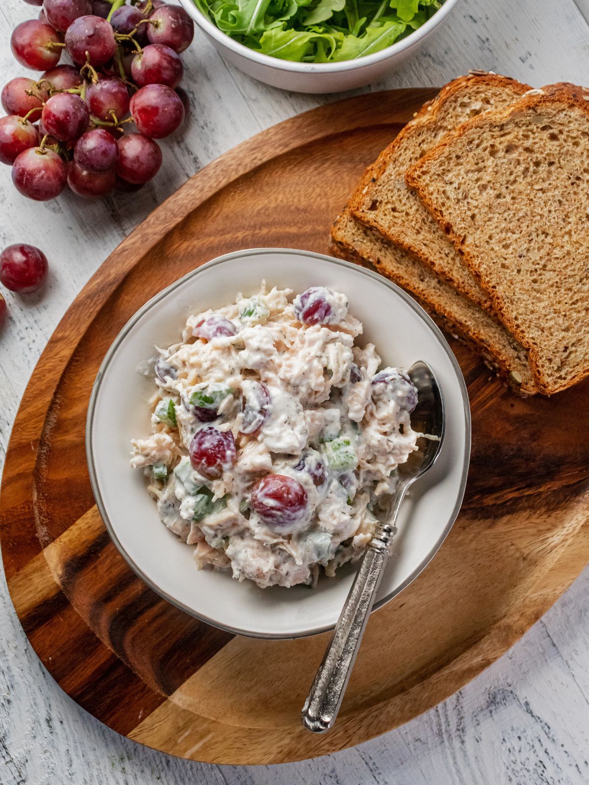 A bowl of high protein chicken salad on a wooden platter with slices of bread to the side and a bunch of grapes.