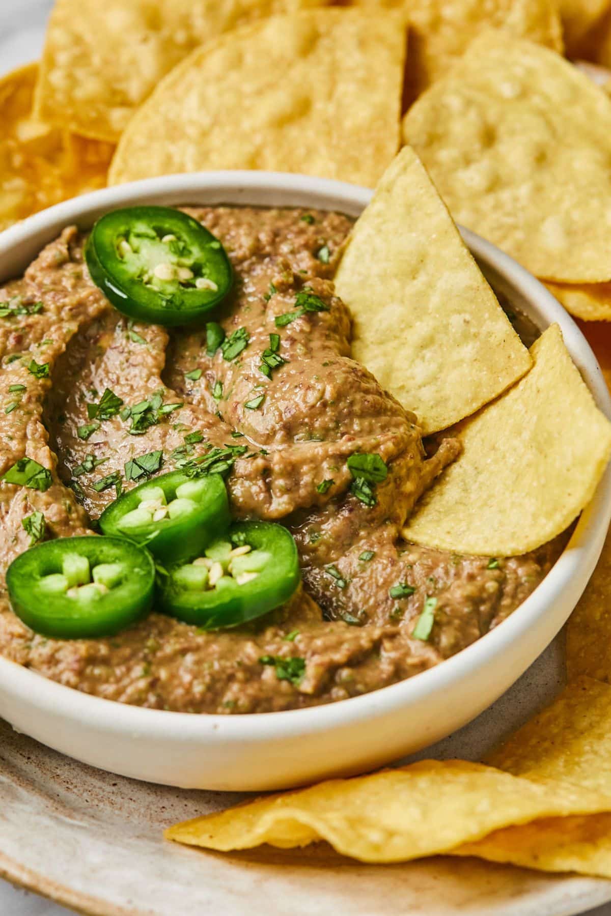 A bowl of spicy black bean hummus without tahini in a bowl with chips dipping in and garnished with cilantro and jalapeno.