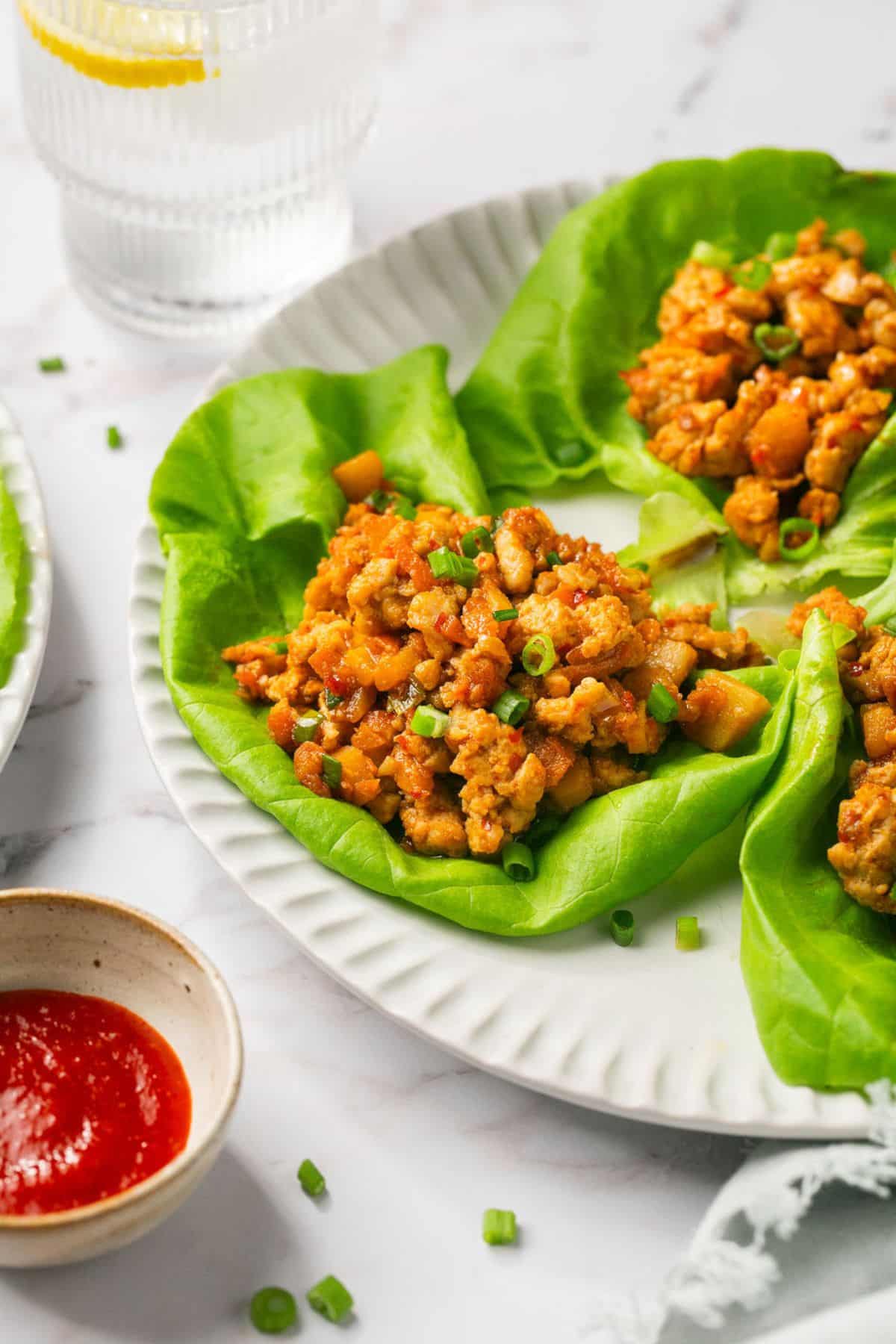 A plate of chicken lettuce wraps with a bowl of chili sauce on the side and sprinkled with green onions. 