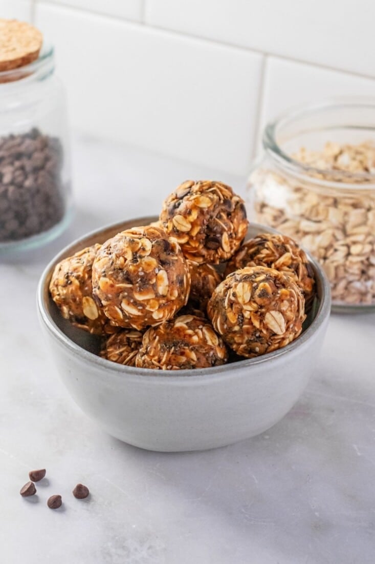 A bowl of almond butter protein balls with a jar of oats and chocolate chips in the background.