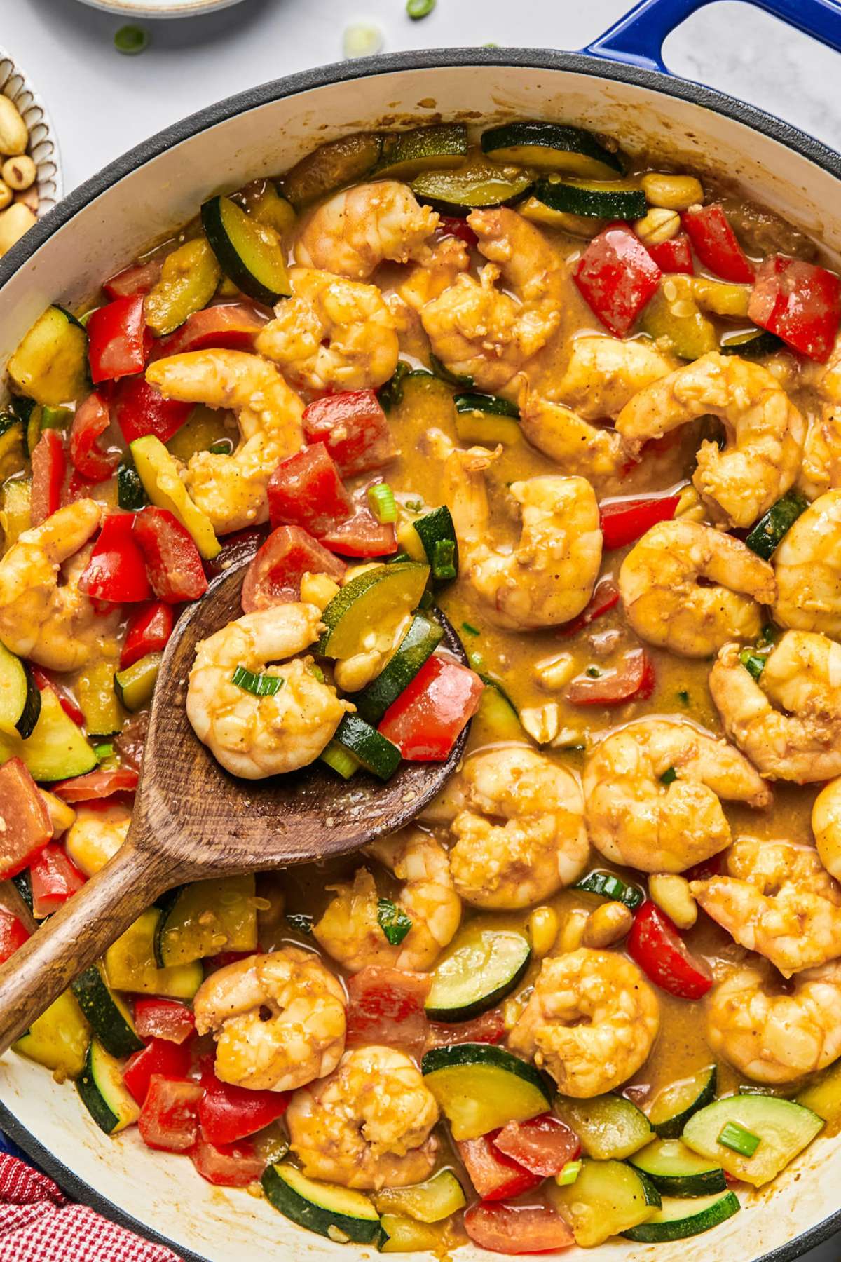 A skillet of kung pao shrimp on a table with a wooden spoon resting on the side with a bit of the shrimp in it.