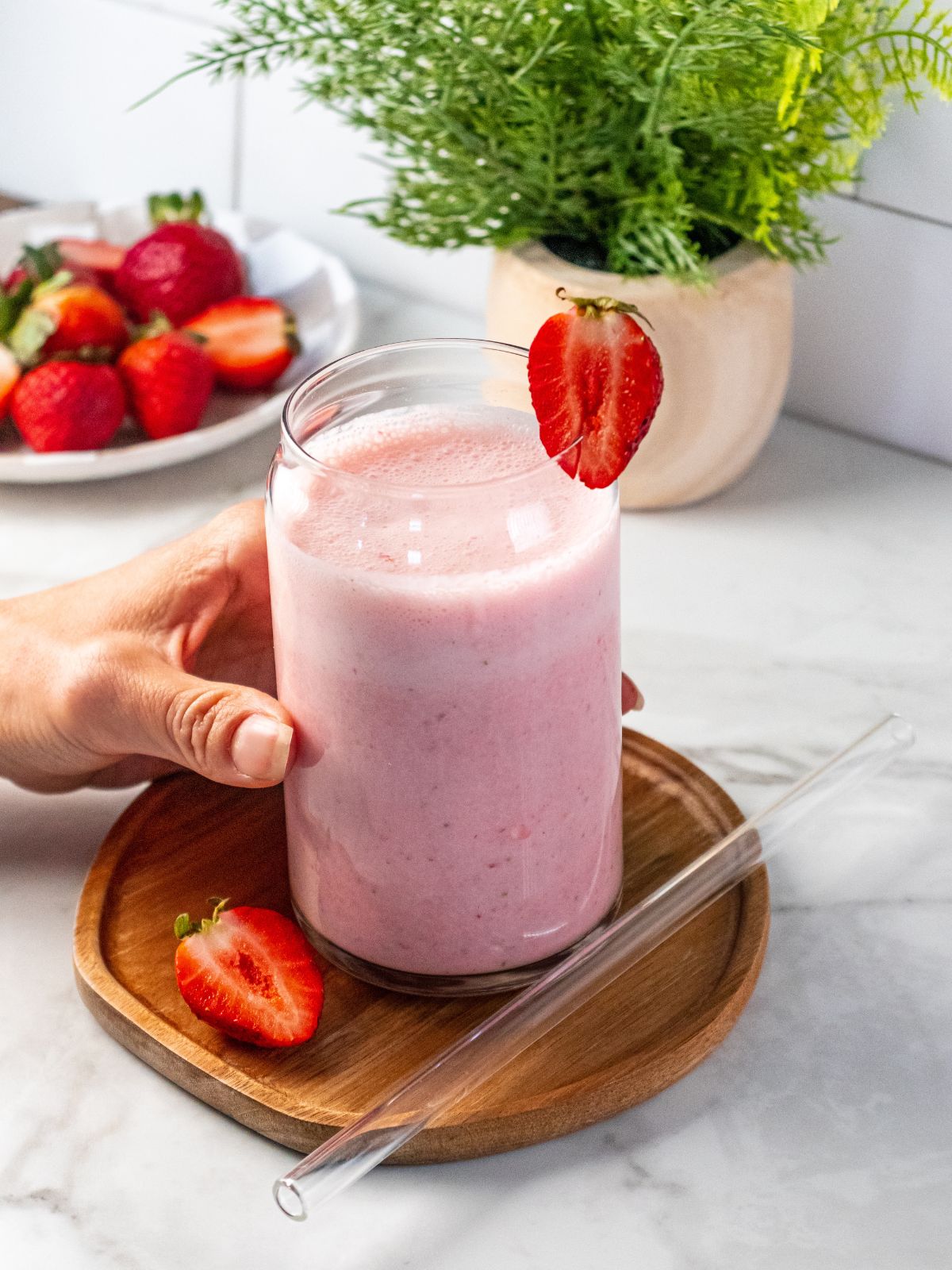A hand reaching in to grab a pink greek yogurt smoothie in a glass with a glass straw on the side and half a strawberry.