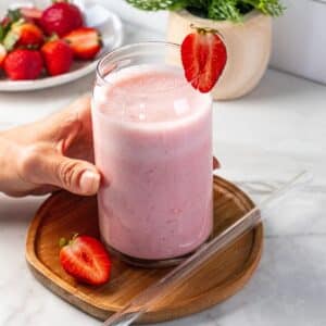 A hand reaching to grab a strawberry greek yogurt smoothie on the table with fresh berries and a glass straw to the side.