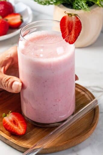 A hand reaching to grab a strawberry greek yogurt smoothie on the table with fresh berries and a glass straw to the side.