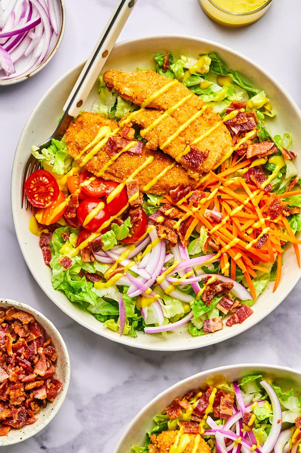 Crispy chicken tender salad drizzled with honey mustard and surrounded by bowls of ingredients. 