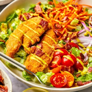 A crispy chicken tender salad with two chicken tenders, tomatoes, and other veggies drizzled with honey mustard over lettuce.