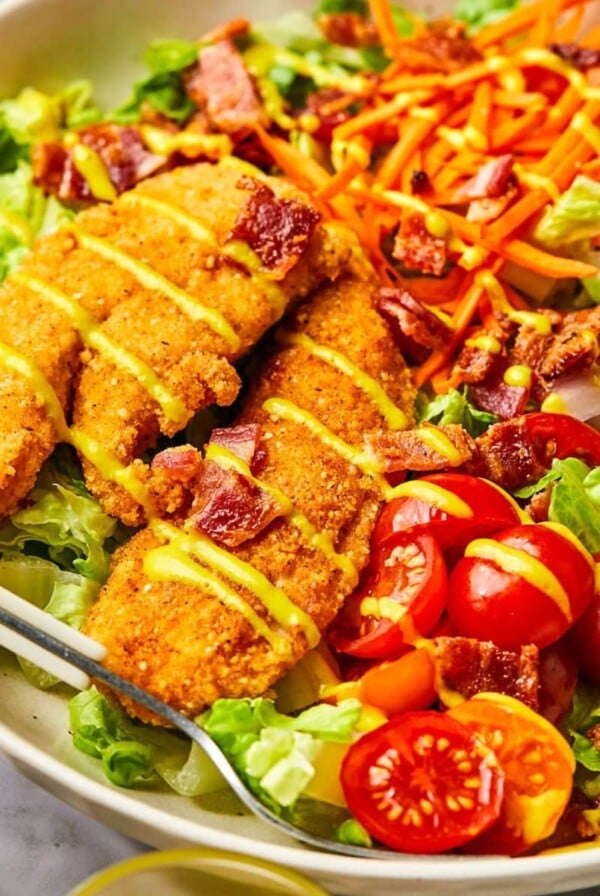 A crispy chicken tender salad with two chicken tenders, tomatoes, and other veggies drizzled with honey mustard over lettuce.