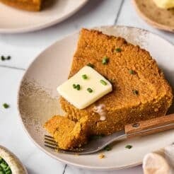 A slice of pumpkin cornbread on a plate with a pat of butter on top and a piece cut off.