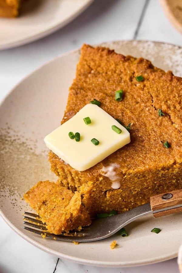 A slice of pumpkin cornbread on a plate with a pat of butter on top and a piece cut off.