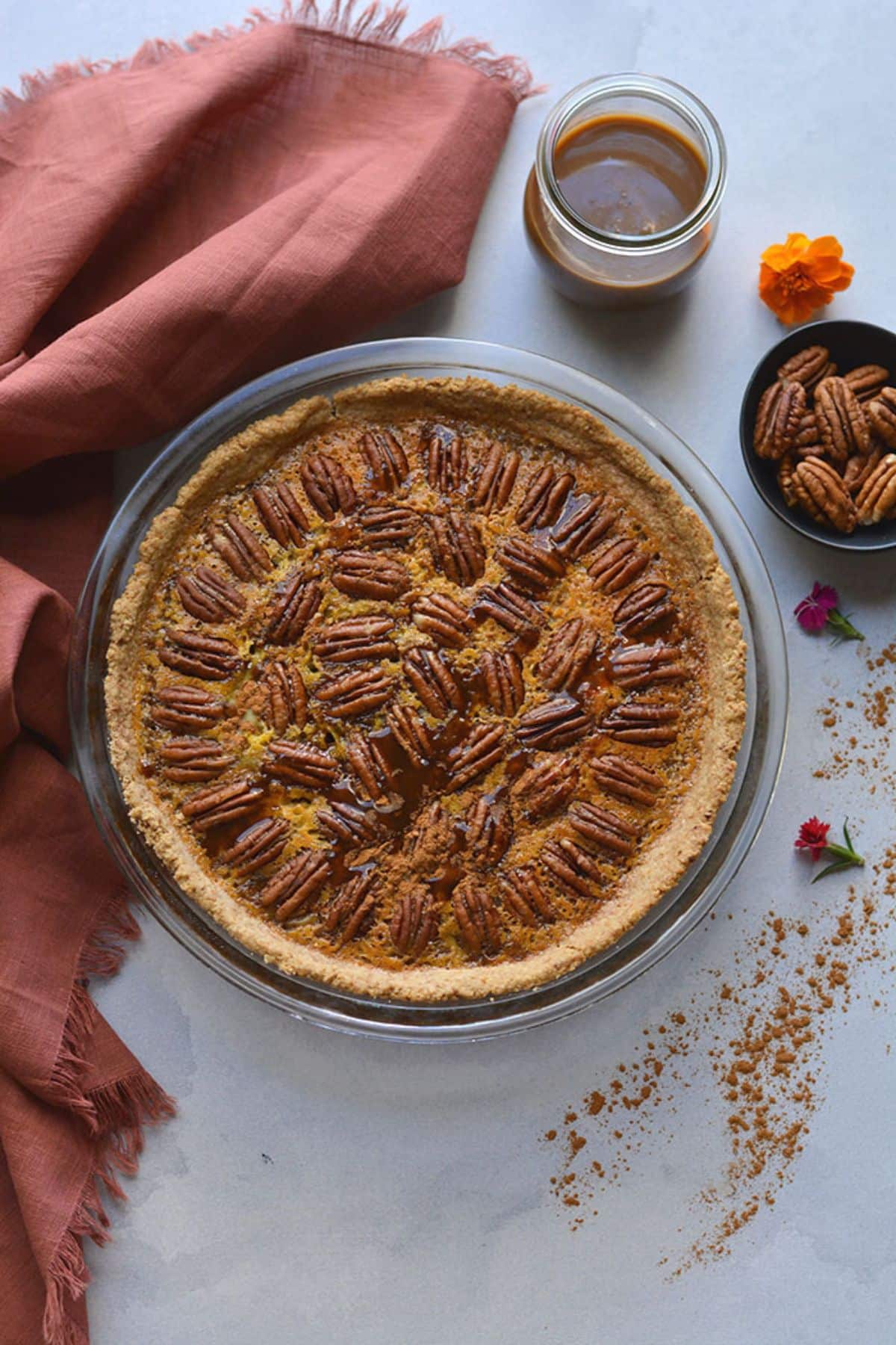 Healthy pecan pie on a table with a jar of caramel sauce and dish of pecans. 
