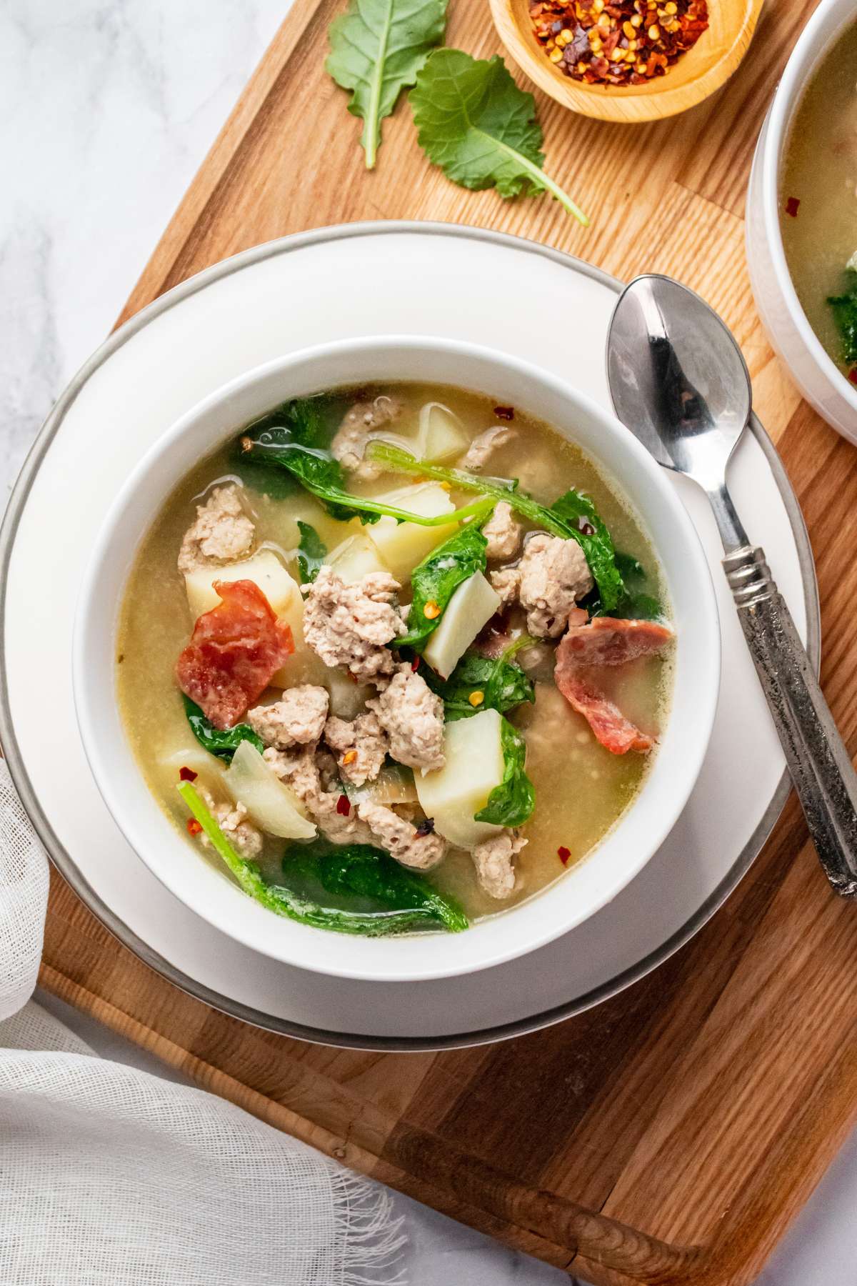 A bowl of skinny zuppa toscana on the table on a cutting board with another bowl in the corner and a linen napkin on the towel.