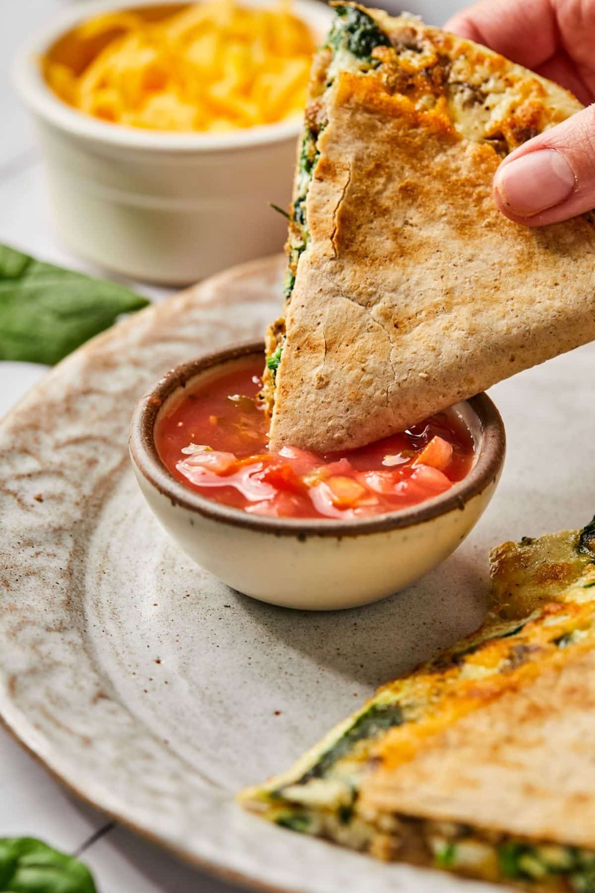 A hand dipping a triangle of breakfast quesadilla into a bowl of salsa.