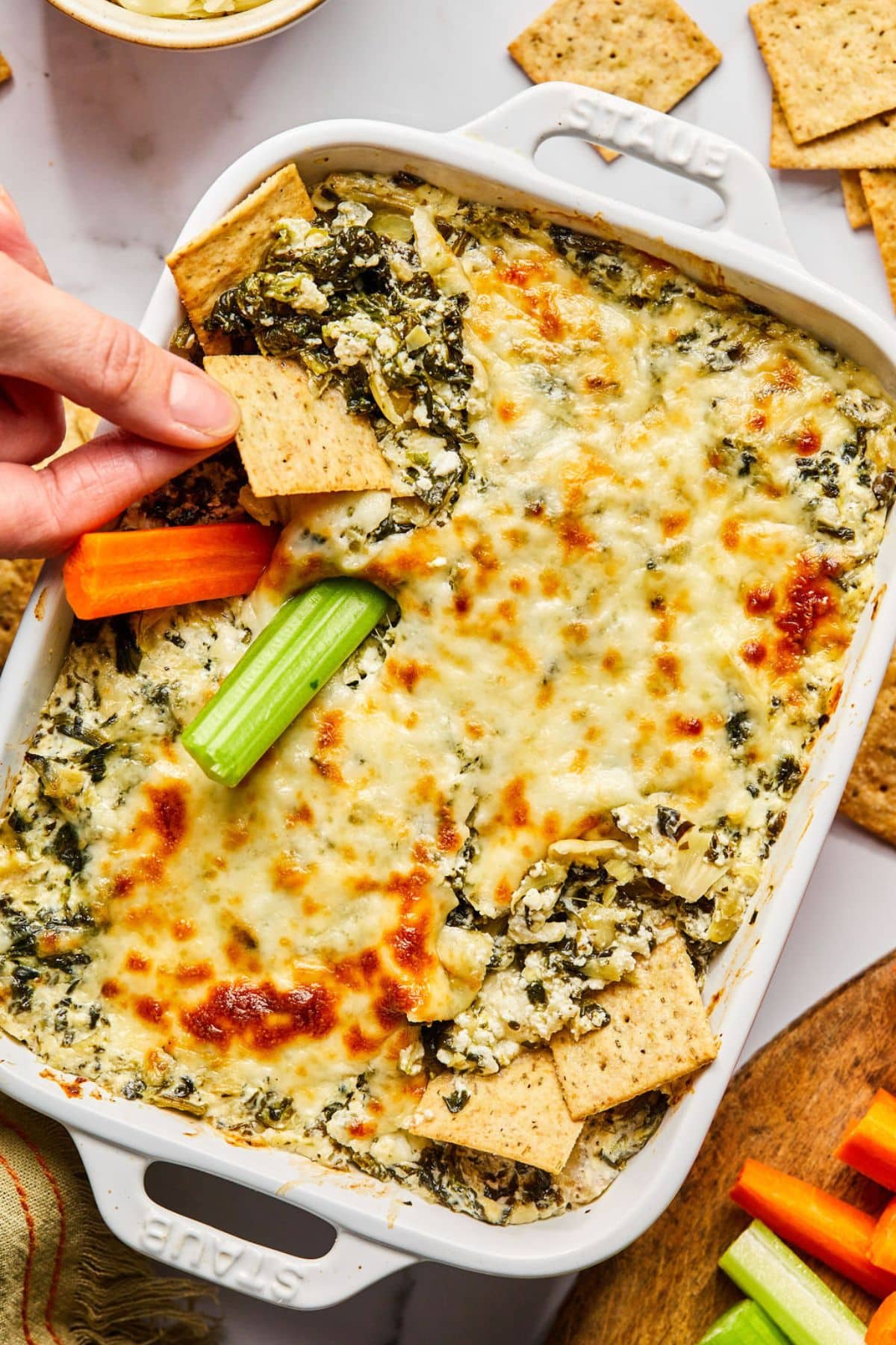 A hand dipping into a cottage spinach artichoke dip in a baking dish with a cracker.