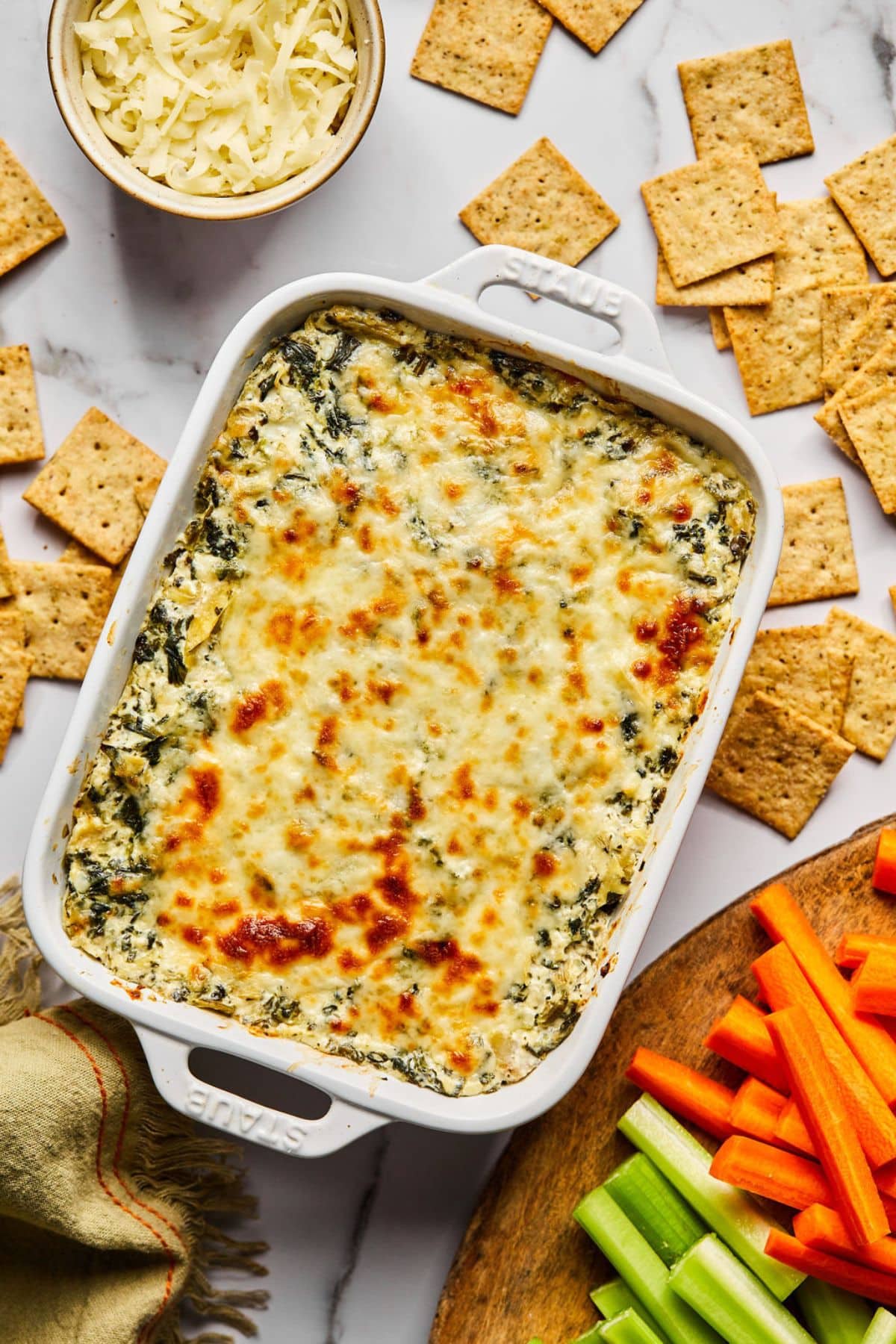 Cottage cheese spinach artichoke dip in a baking dish with crackers scattered around and a bowl of cheese.