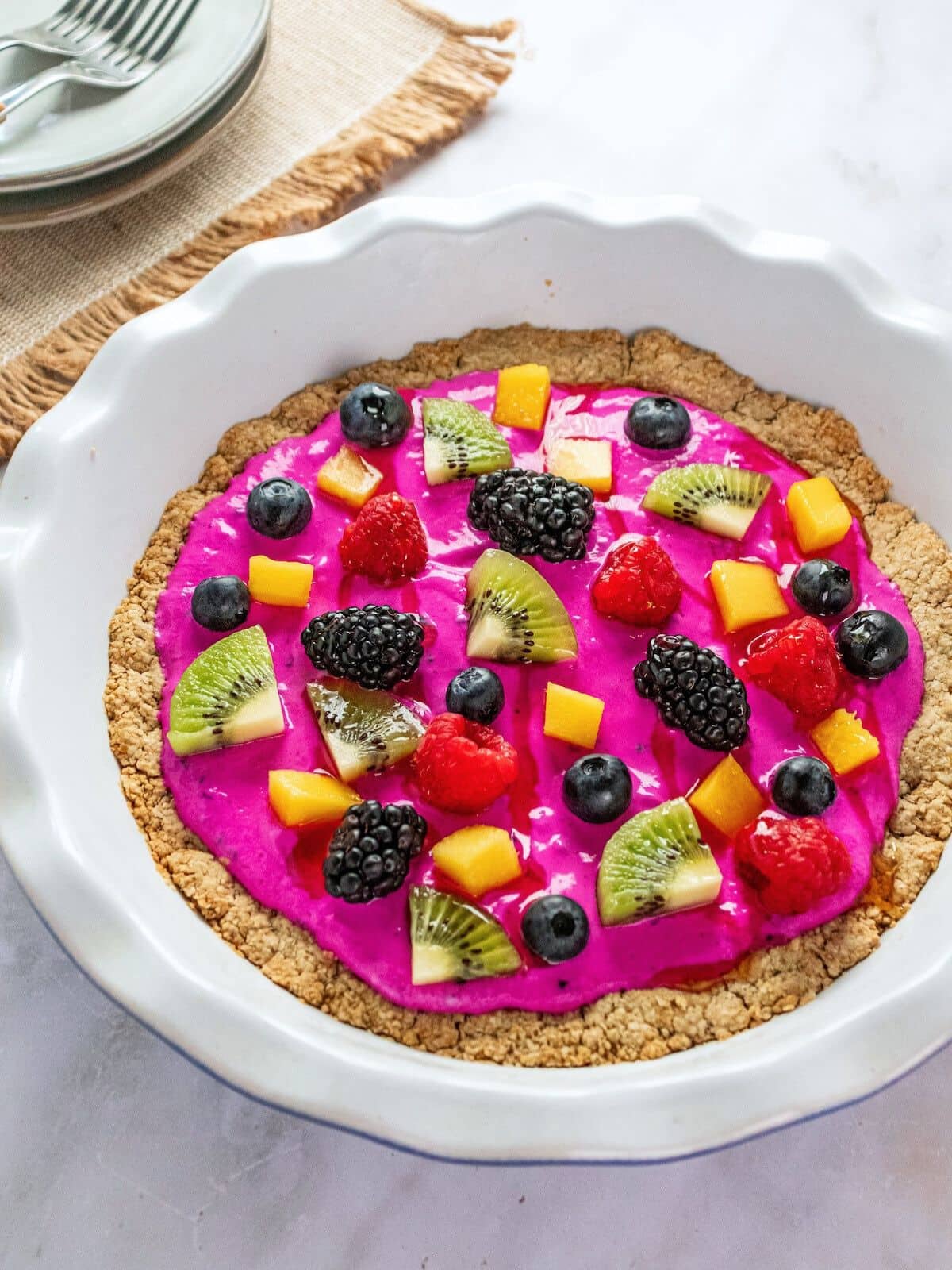 Greek yogurt fruit pizza in a white pie dish topped with fresh fruit and bright pink yogurt.
