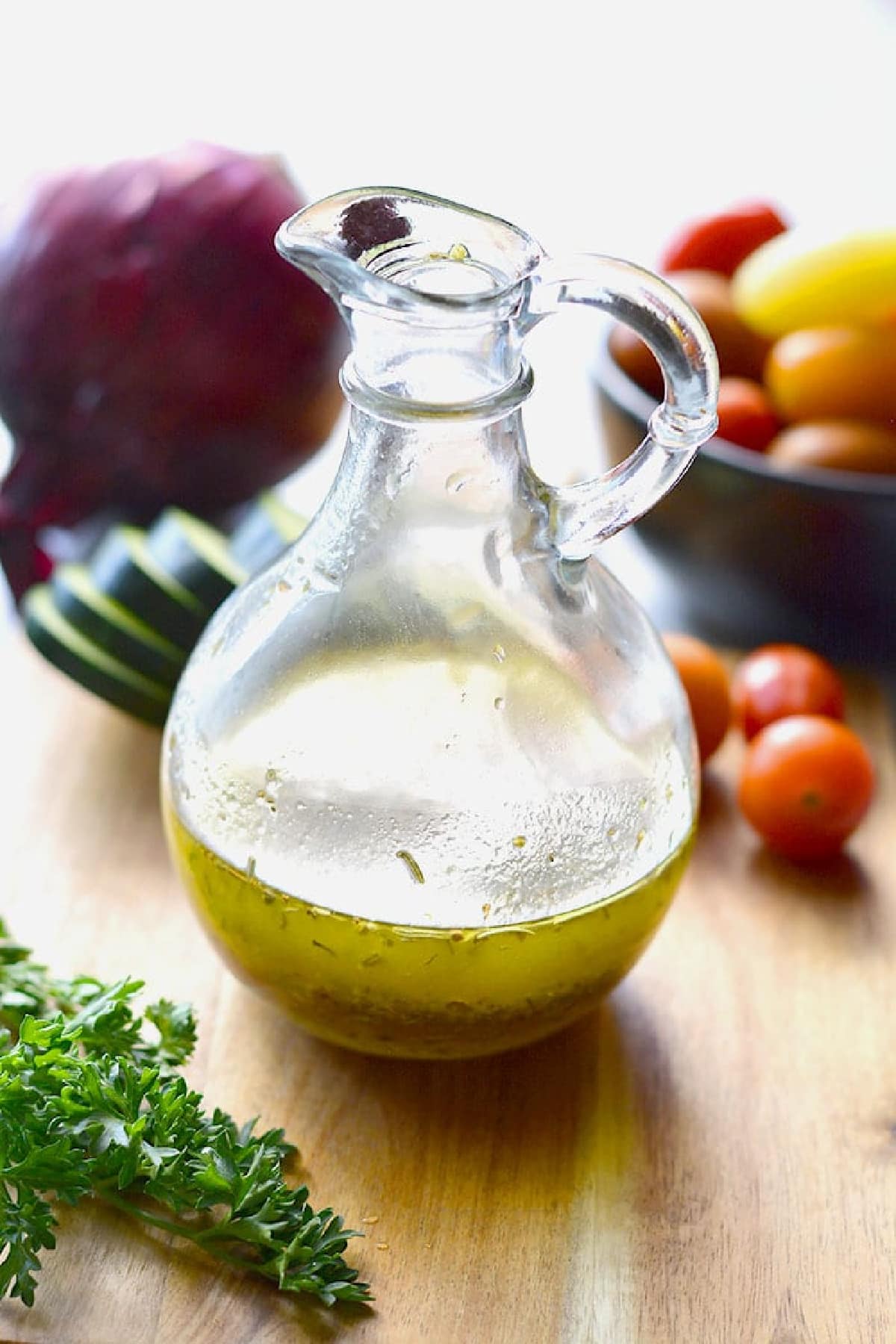 dressing in a clear dressing bottle on a wooden cutting board with vegetables behind it.