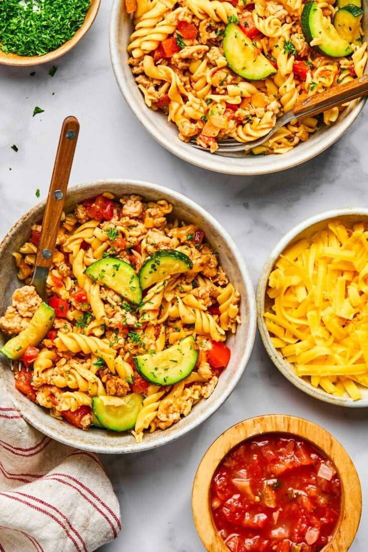 Bowls of healthy taco pasta on a table with bowls of salsa and cheese too.