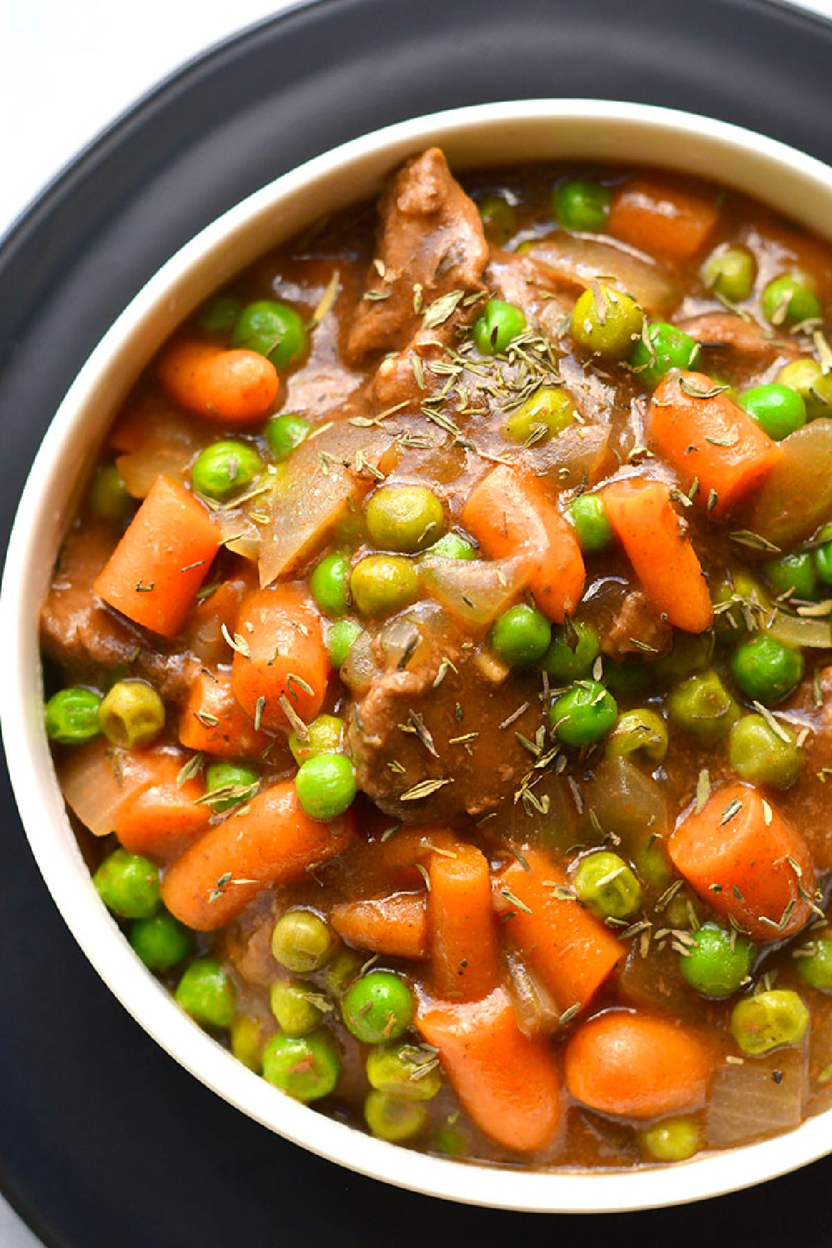 Bowl of healthy beef stew on a plate with chunks of beef, carrots, and peas.