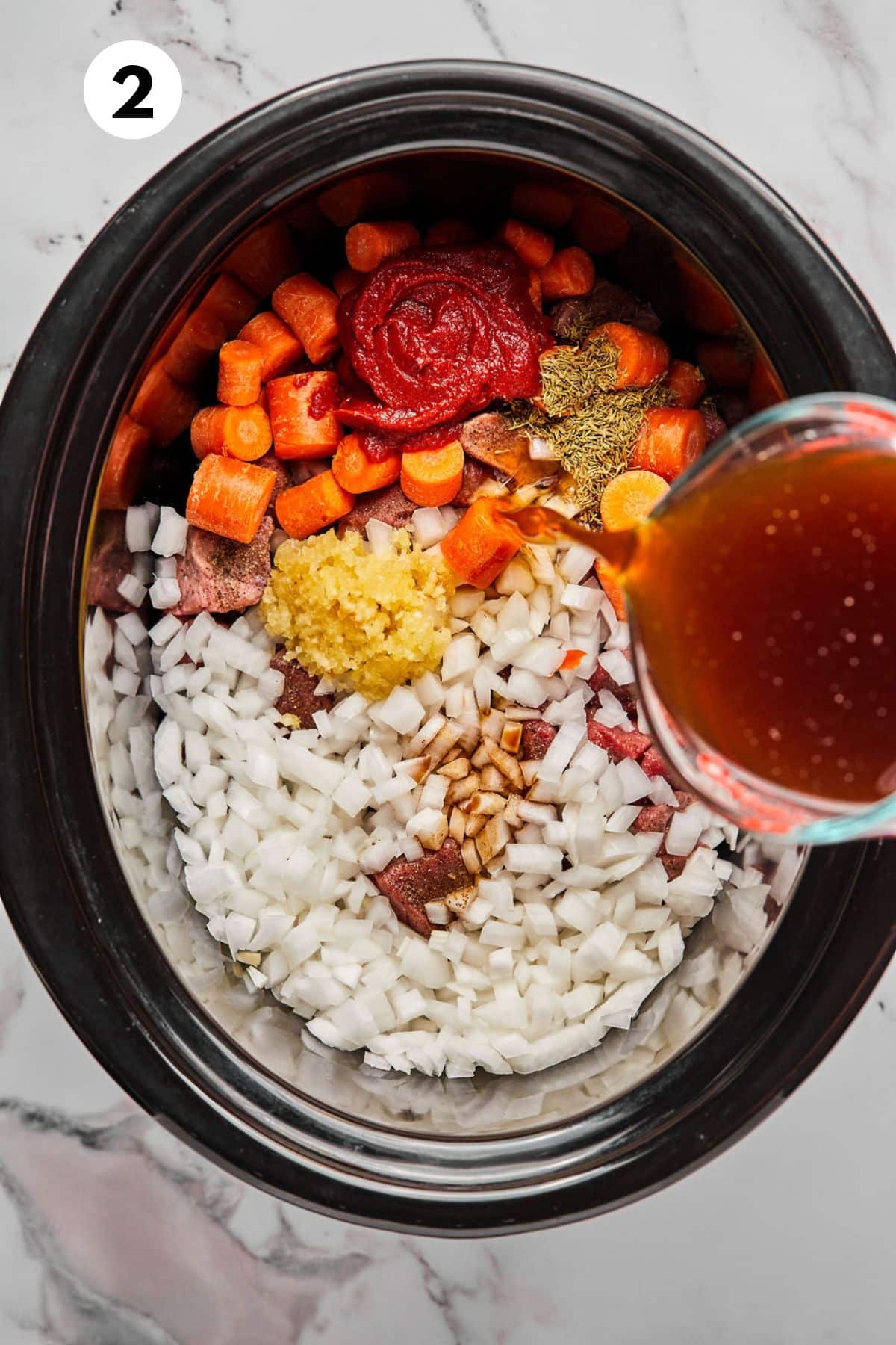 Vegetables and seasonings added to the beef stew meat while beef broth is pouring from a cup.