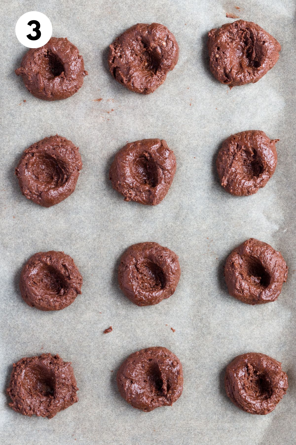Chocolate thumbprint cookie dough balls on a baking sheet with a thumbprint in the middle of each.